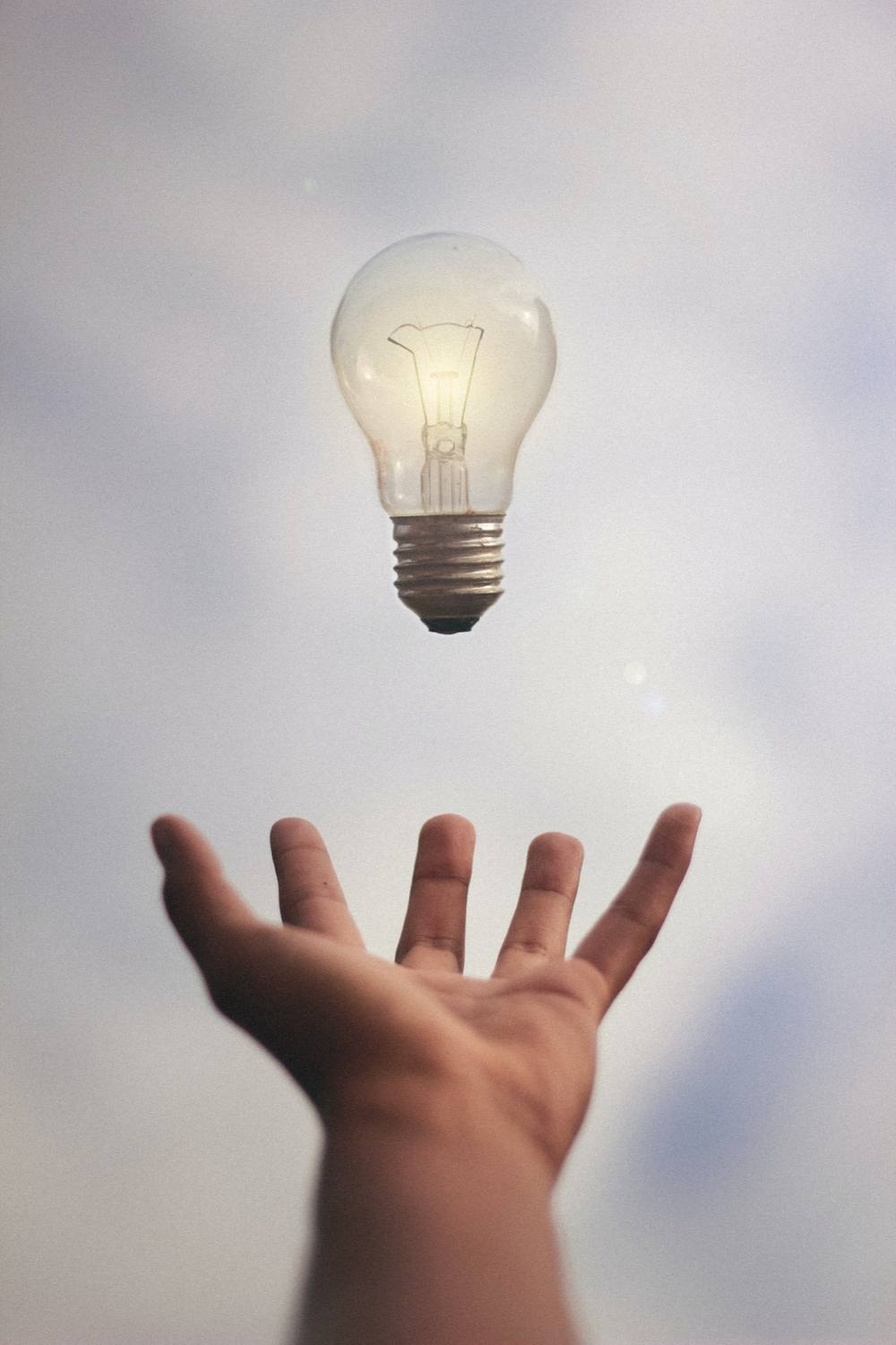 An open hand reaching up toward a lit incandescent bulb suspended in a pale sky